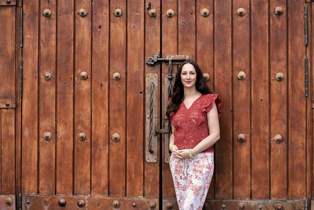 J stands beside a large wooden door in a red blouse and floral skirt.